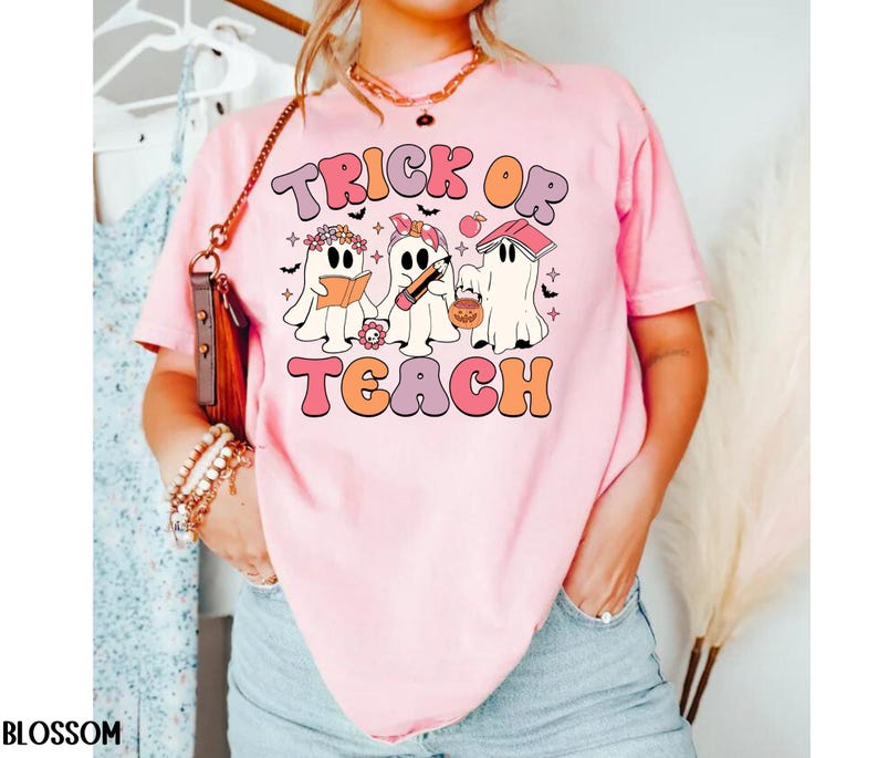 May include: Pink t-shirt with the words "Trick or Teach" in a retro font. The graphic features three cartoon ghosts, one reading a book, one with a pencil, and one with a jack-o'-lantern bucket. The shirt is a comfortable, casual style.