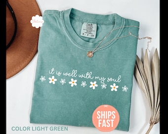 May include: Light green t-shirt with the phrase "it is well with my soul" in white script and daisy graphics. A gold necklace with a pendant rests on the shirt. The shirt is folded and displayed with a brown hat and dried plant.