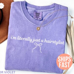 May include: A lilac t-shirt with the white text "I'm literally just a hairstylist". A gold necklace with a pendant is on the shirt. The shirt has a crew neck and a small bow graphic.