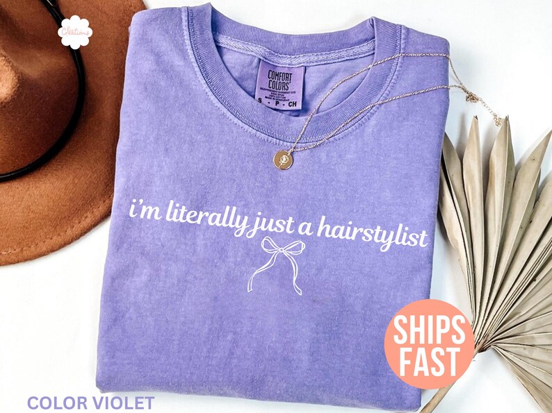 May include: A lilac t-shirt with the white text "I'm literally just a hairstylist". A gold necklace with a pendant is on the shirt. The shirt has a crew neck and a small bow graphic.