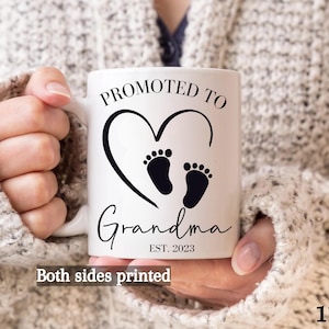 K&ouml;nnte beinhalten: Wei&szlig;e Keramiktasse mit schwarzem Text und einem Herzmotiv mit zwei Fu&szlig;abdr&uuml;cken. Der Text lautet "Bef&ouml;rdert zur Oma Est. 2023". Die Tasse ist 11 Unzen.