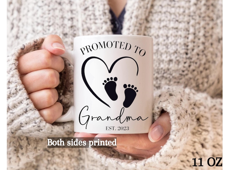K&ouml;nnte beinhalten: Wei&szlig;e Keramiktasse mit schwarzem Text und einem Herzmotiv mit zwei Fu&szlig;abdr&uuml;cken. Der Text lautet "Bef&ouml;rdert zur Oma Est. 2023". Die Tasse ist 11 Unzen.