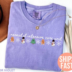 May include: A lavender Comfort Colors t-shirt with the words "neonatal intensive care unit" in white script. The shirt features gingerbread men, snowmen, Christmas trees, and snowflakes. A gold necklace is draped on the shirt.