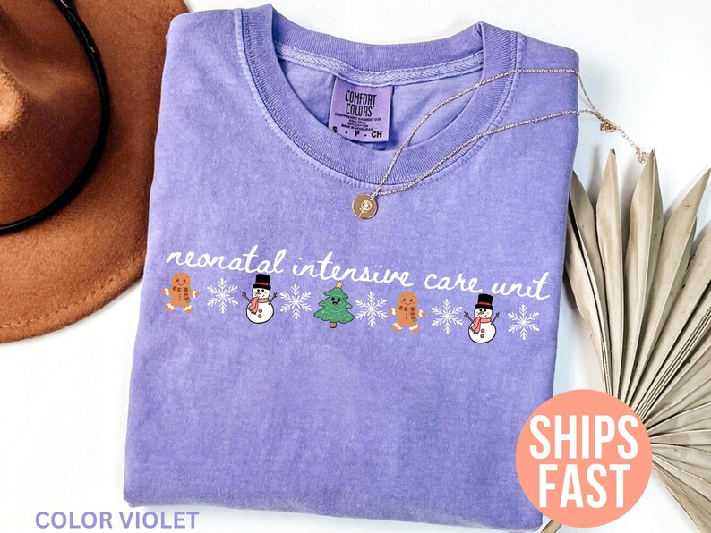 May include: A lavender Comfort Colors t-shirt with the words "neonatal intensive care unit" in white script. The shirt features gingerbread men, snowmen, Christmas trees, and snowflakes. A gold necklace is draped on the shirt.