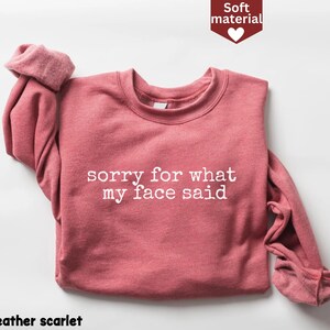 May include: A heather scarlet sweatshirt with the text "sorry for what my face said" in white. The long-sleeved top is made of a soft material and has a crew neck. The image also includes a small graphic that says "Soft material".