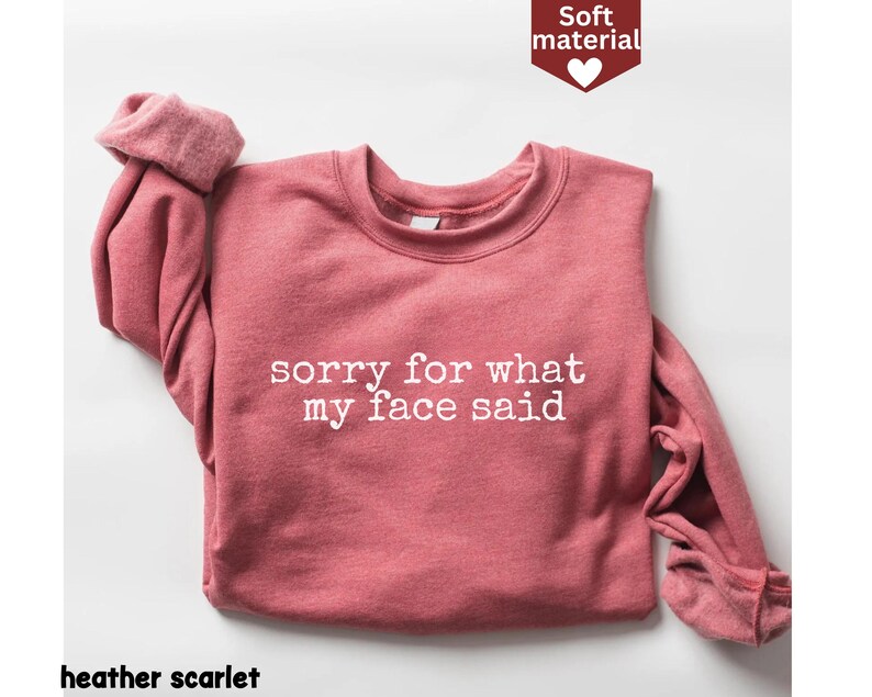 May include: A heather scarlet sweatshirt with the text "sorry for what my face said" in white. The long-sleeved top is made of a soft material and has a crew neck. The image also includes a small graphic that says "Soft material".