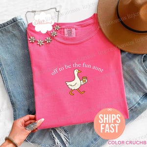 May include: A bright pink short-sleeved t-shirt featuring a graphic of a white goose holding a coffee cup with the text off to be the fun aunt printed above it. The shirt is paired with blue denim jeans, a brown hat, and a floral necklace.