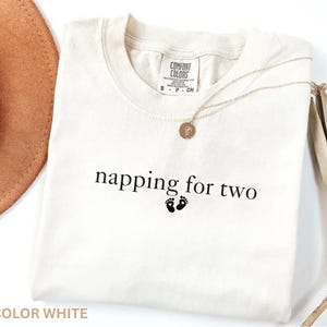 Puede incluir: Camiseta blanca con el texto "napping for two" y dos peque&ntilde;as huellas negras. Un collar dorado con un colgante est&aacute; sobre la camiseta. La camiseta est&aacute; etiquetada como "Comfort Colors".