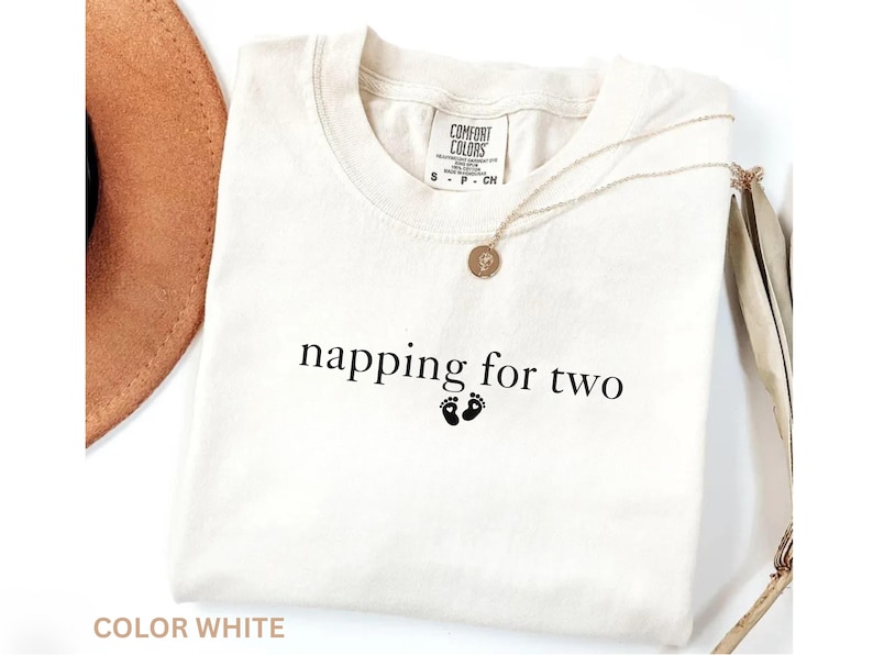 Puede incluir: Camiseta blanca con el texto "napping for two" y dos peque&ntilde;as huellas negras. Un collar dorado con un colgante est&aacute; sobre la camiseta. La camiseta est&aacute; etiquetada como "Comfort Colors".