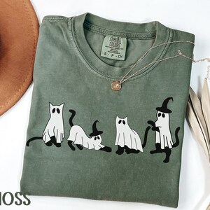 May include: Moss green t-shirt with a graphic of four cartoon cats dressed as ghosts, some wearing witch hats. The shirt has a crew neck and short sleeves. A gold necklace with a pendant is draped on the shirt. The word "MOSS" is printed at the bottom.