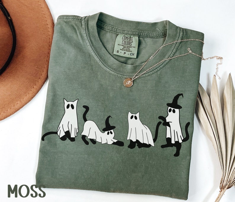 May include: Moss green t-shirt with a graphic of four cartoon cats dressed as ghosts, some wearing witch hats. The shirt has a crew neck and short sleeves. A gold necklace with a pendant is draped on the shirt. The word "MOSS" is printed at the bottom.