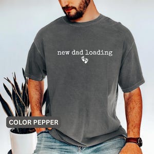 Pode incluir: Camiseta cor pimenta com o texto branco "new dad loading" e pequenas pegadas brancas. A camisa tem um ajuste folgado e parece ser feita de material macio. Uma peça de roupa casual.