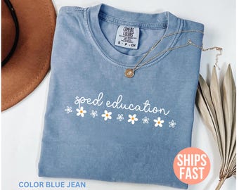 Puede incluir: Una camiseta azul vaquero con las palabras "sped education" en escritura blanca y detalles de margaritas. Un collar de oro con un colgante descansa sobre la camisa. La camisa tiene una etiqueta Comfort Colors.