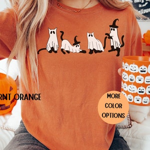 May include: Burnt orange Halloween-themed sweatshirt with a graphic of four cartoon cats and ghosts. The cats and ghosts are wearing witch hats and have black tails. The text "Burnt Orange" is at the bottom left, and "More Color Options" is in a circle.