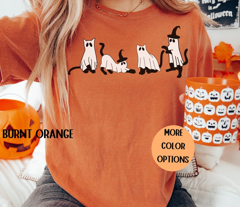 May include: Burnt orange Halloween-themed sweatshirt with a graphic of four cartoon cats and ghosts. The cats and ghosts are wearing witch hats and have black tails. The text "Burnt Orange" is at the bottom left, and "More Color Options" is in a circle.