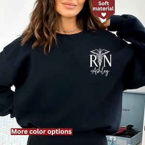 Könnte beinhalten: Schwarzer Pullover mit einem weißen Aufdruck eines Caduceus und den Buchstaben "RN" mit dem Namen "Ashley" darunter. Der Pullover ist aus weichem Material.