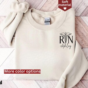 May include: A cream-colored sweatshirt with a black graphic of a medical caduceus and the text "RN Ashley".