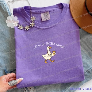 Peut inclure: Un t-shirt violet Comfort Colors avec le texte "off to do BCBA things" et un canard de dessin animé tenant une tasse de café. Le t-shirt est de taille L-G-G.