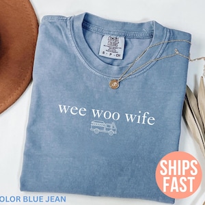 May include: Blue jean colored t-shirt with the words "wee woo wife" and a fire truck graphic. A gold necklace with a pendant rests on the shirt. The shirt is folded, and a brown hat is partially visible.