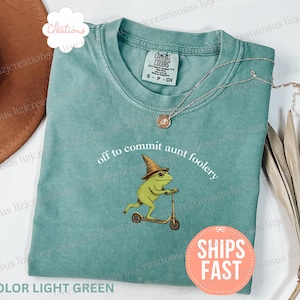May include: Light green t-shirt with the phrase "off to commit aunt foolery" and a cartoon frog wearing a witch hat riding a scooter. A silver necklace with a pendant is on the shirt.