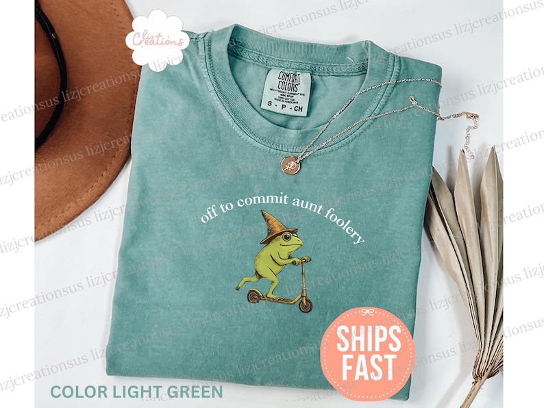 May include: Light green t-shirt with the phrase "off to commit aunt foolery" and a cartoon frog wearing a witch hat riding a scooter. A silver necklace with a pendant is on the shirt.
