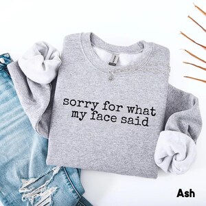 May include: A light gray sweatshirt with the text "sorry for what my face said" in black. The sweatshirt is paired with light blue denim jeans. The sleeves are cuffed, revealing a white interior. The word "Ash" is at the bottom.