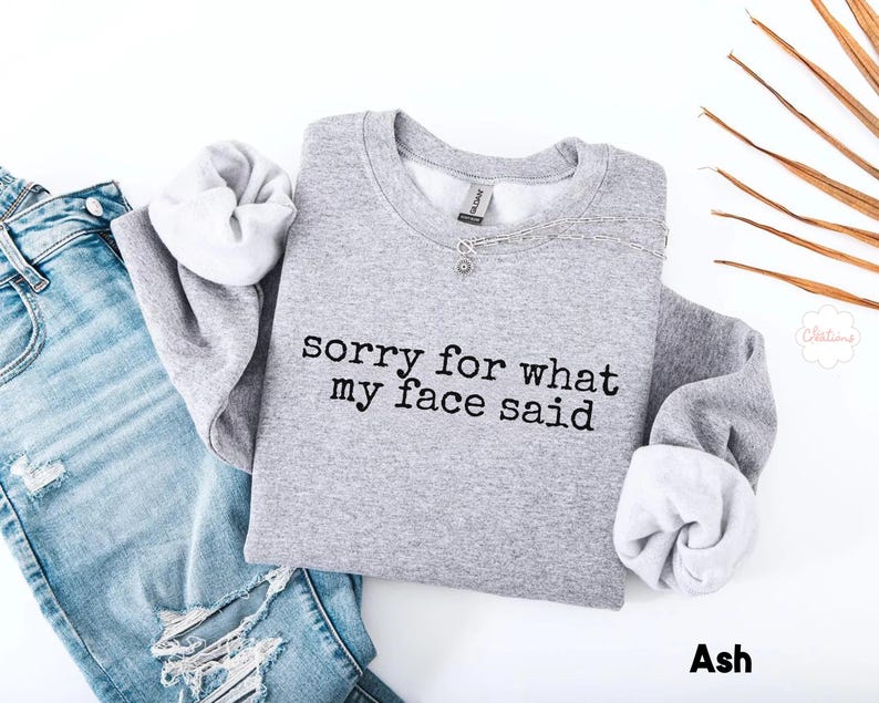 May include: A light gray sweatshirt with the text "sorry for what my face said" in black. The sweatshirt is paired with light blue denim jeans. The sleeves are cuffed, revealing a white interior. The word "Ash" is at the bottom.