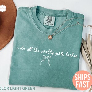 May include: Light green t-shirt with the text "i do all the pretty girls lashes" and a bow graphic. A gold necklace with a pendant rests on the shirt. The shirt is a size S-P-CH.