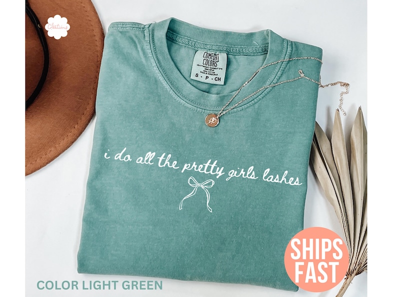 May include: Light green t-shirt with the text "i do all the pretty girls lashes" and a bow graphic. A gold necklace with a pendant rests on the shirt. The shirt is a size S-P-CH.