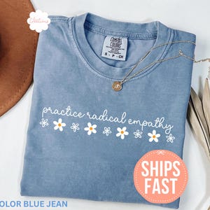 May include: A blue jean colored t-shirt with the phrase "practice radical empathy" in white script and daisy graphics. A gold necklace with a pendant rests on the shirt. The shirt is folded and displayed with a brown hat and dried plant.