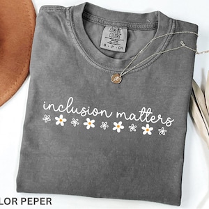 May include: A gray t-shirt with the words "inclusion matters" in white script, decorated with small daisy graphics. A silver necklace with a pendant rests on the shirt. The shirt is folded and has a tag.
