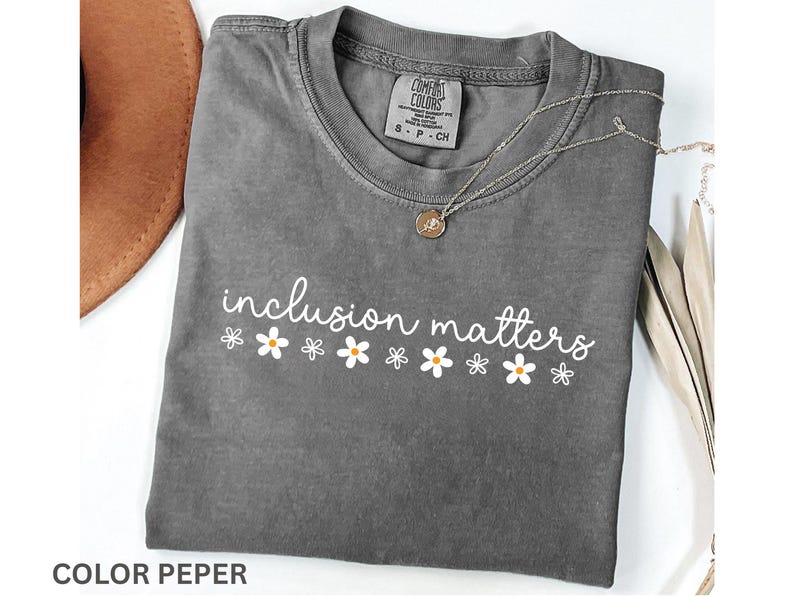 May include: A gray t-shirt with the words "inclusion matters" in white script, decorated with small daisy graphics. A silver necklace with a pendant rests on the shirt. The shirt is folded and has a tag.