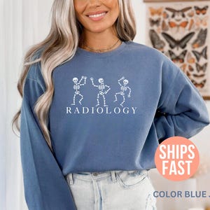 May include: Blue jean-colored sweatshirt with a white graphic of three dancing skeletons above the word "RADIOLOGY". The sweatshirt has long sleeves and a crew neck. The image also includes the text "SHIPS FAST" and "COLOR BLUE JEAN".