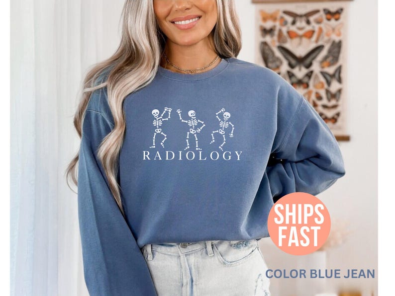 May include: Blue jean-colored sweatshirt with a white graphic of three dancing skeletons above the word "RADIOLOGY". The sweatshirt has long sleeves and a crew neck. The image also includes the text "SHIPS FAST" and "COLOR BLUE JEAN".
