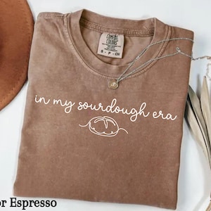 May include: A brown t-shirt with the text "in my sourdough era" and a simple line drawing of a loaf of bread. The shirt is a comfortable, casual style.