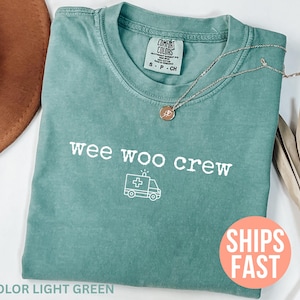 Puede incluir: Camiseta verde claro con el texto "wee woo crew" y un gráfico de ambulancia. Un collar plateado con un colgante descansa sobre la camisa. La camisa está doblada y colocada sobre una superficie con un sombrero marrón.