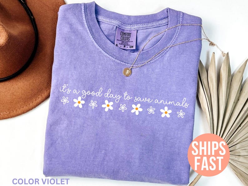 K&ouml;nnte beinhalten: Ein violettes T-Shirt mit dem wei&szlig;en Schriftzug "it's a good day to save animals" und wei&szlig;en G&auml;nsebl&uuml;mchen-Grafiken. Eine goldene Halskette liegt auf dem Shirt. Das Shirt ist mit "Comfort Colors" gekennzeichnet.