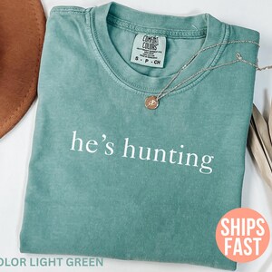 May include: Light green t-shirt with the phrase "he's hunting" in white. A gold-colored necklace with a pendant rests on the shirt. A brown hat and dried plant stems are also visible.