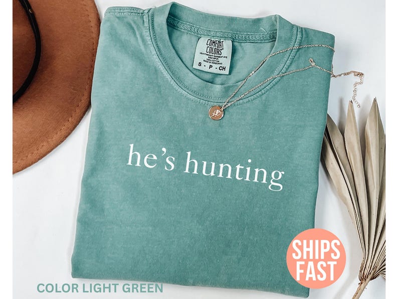 May include: Light green t-shirt with the phrase "he's hunting" in white. A gold-colored necklace with a pendant rests on the shirt. A brown hat and dried plant stems are also visible.