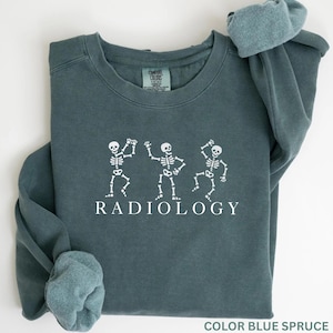 May include: Blue spruce-colored sweatshirt with white skeleton graphics dancing. The word "RADIOLOGY" is printed in white below the skeletons. The sweatshirt has a crew neck and long sleeves.