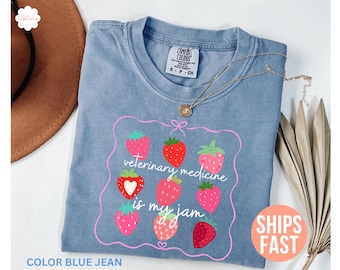 May include: Blue jean colored t-shirt with a pink framed graphic of strawberries and the text "veterinary medicine is my jam". A gold necklace is draped across the shirt. The shirt has a Comfort Colors tag.