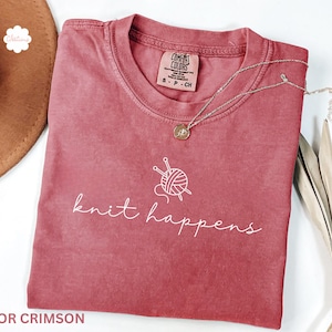 May include: Crimson-colored t-shirt with the words "knit happens" in white script, along with a graphic of yarn and knitting needles. A silver necklace with a pendant rests on the shirt. The shirt is folded.