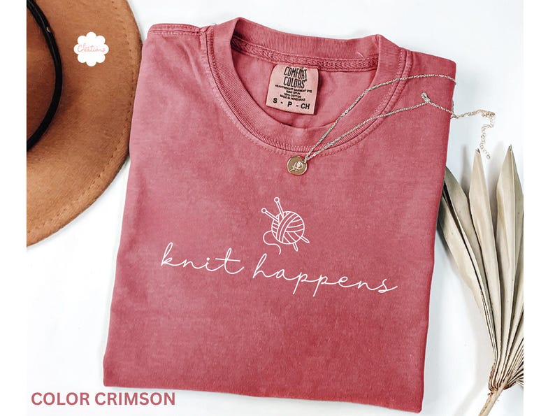 May include: Crimson-colored t-shirt with the words "knit happens" in white script, along with a graphic of yarn and knitting needles. A silver necklace with a pendant rests on the shirt. The shirt is folded.