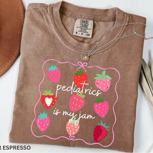 May include: Espresso-coloured t-shirt with a strawberry graphic and the text "pediatrics is my jam." The design features pink and red strawberries inside a pink outline. A silver necklace with a pendant is draped on the shirt.