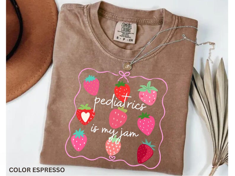 May include: Espresso-coloured t-shirt with a strawberry graphic and the text "pediatrics is my jam." The design features pink and red strawberries inside a pink outline. A silver necklace with a pendant is draped on the shirt.