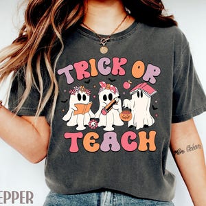 May include: Dark gray t-shirt with the words "TRICK OR TEACH" in a retro font. The shirt features three cartoon ghosts, one reading a book, one holding a pencil, and one with a jack-o'-lantern. The design is in shades of pink, orange, and purple.