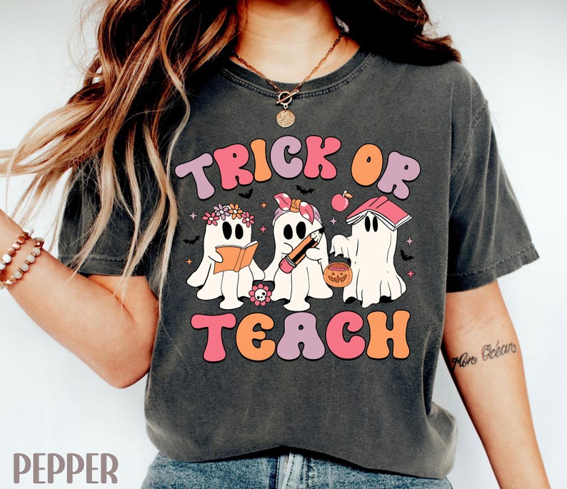 May include: Dark gray t-shirt with the words "TRICK OR TEACH" in a retro font. The shirt features three cartoon ghosts, one reading a book, one holding a pencil, and one with a jack-o'-lantern. The design is in shades of pink, orange, and purple.