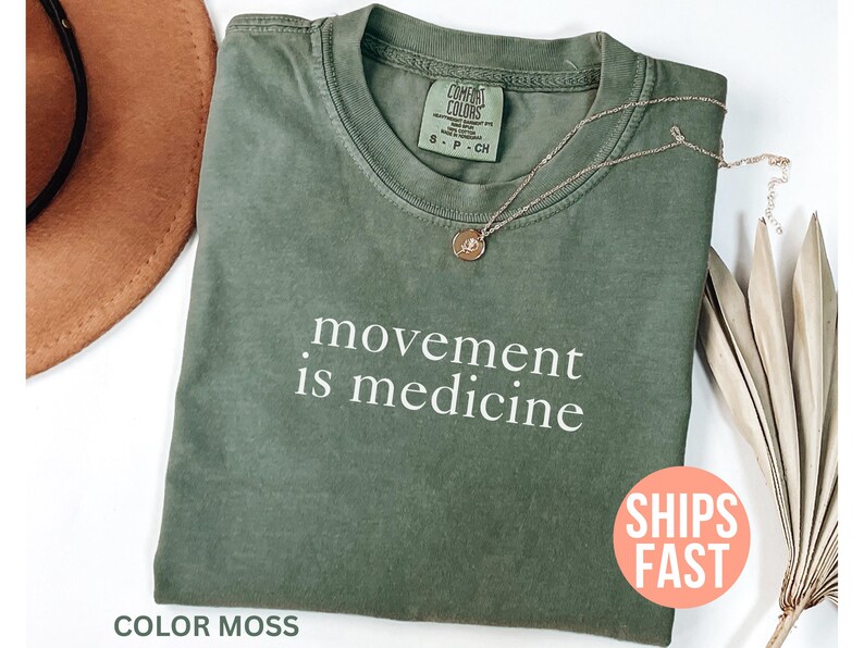 K&ouml;nnte beinhalten: Ein moosgr&uuml;nes T-Shirt mit dem wei&szlig;en Text "movement is medicine". Eine goldfarbene Halskette mit Anh&auml;nger liegt auf dem Shirt. Das Shirt ist gefaltet, und ein brauner Hut ist teilweise sichtbar.