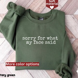 May include: Military green sweatshirt with the text "sorry for what my face said" in white. The sweatshirt has a crew neck and long sleeves. The words "Soft material" and "More color options" are also visible.