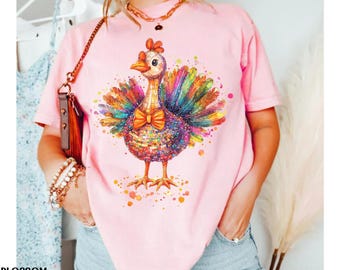 Könnte beinhalten: Hellrosa T-Shirt mit einem Aquarell-Truthahn-Design. Der Truthahn hat eine Fliege und bunte Federn in Blau, Grün, Gelb und Rot. Das Wort "BLOSSOM" ist unten links aufgedruckt.